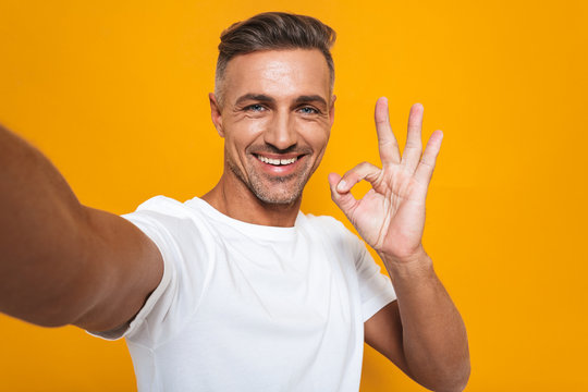 Image of brunette man 30s in white t-shirt smiling and taking selfie photo