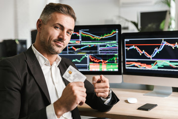 Portrait of european businessman 30s wearing suit holding credit card while sitting in office