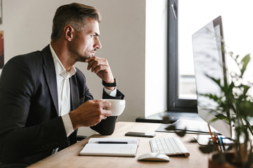 Image of european businessman drinking coffee while working on computer in office