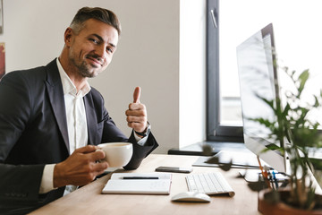 Image of joyous businessman drinking coffee while working on computer in office