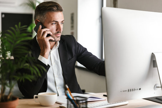 Image of smart businessman talking on cell phone while working on computer in office