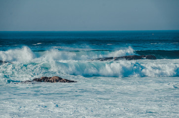 Ocean seascape during the storm with blue waves with splashes and foam over the stones in sunny weather