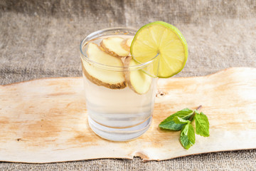 Fototapeta premium Glass with ginger water and lime slice and mint on piece of bark on burlap. 