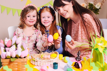 young woman mother with little girls daughters preparing for the celebration of Easter and coloring eggs sitting at the table on the background of the Easter decor