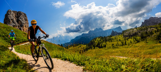 Naklejka na ściany i meble Cycling woman and man riding on bikes in Dolomites mountains landscape. Couple cycling MTB enduro trail track. Outdoor sport activity.