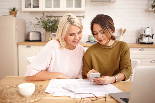 Indoor image of beautiful dark haired young woman holding mobile phone, showing her attractive middle aged mother how to pay mortgage or apartment rent online using application on electronic gadget