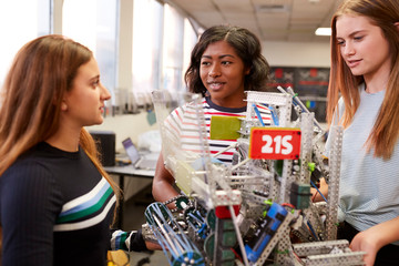 Female University Students Carrying Machine In Science Robotics Or Engineering Class