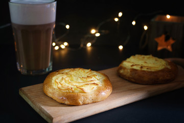 Two cheesecakes with cottage cheese lie on a wooden board. In the background, a glass of coffee and lights of garland in blur.