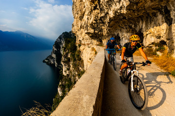 Cycling woman and man riding on bikes at sunrise mountains and Garda lake landscape. Couple cycling MTB enduro flow sentiero ponale trail track. Outdoor sport activity.