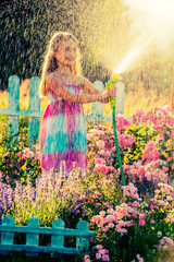 Playfull girl watering flowers with rain in the garden at summer day. Child using garden hose on sunny day at sunset lite. Little gardener playing in garden.