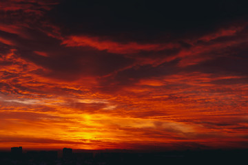 Cityscape with fiery blood vampire dawn. Amazing warm dramatic cloudy sky above dark silhouettes of city buildings. Orange sunlight. Atmospheric background of sunrise in overcast weather. Copy space.
