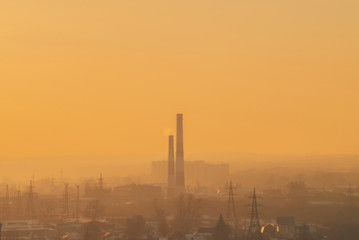 Obraz premium Smog among silhouettes of buildings on sunrise. Smokestack in dawn sky. Environmental pollution on sunset. Harmful fumes from stack above city. Mist urban background with warm orange yellow sky.