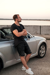 Gorgeous sexy man on a vacation car trip in summer day. Luxury grey sport car. Man in casual outfit. Sunset