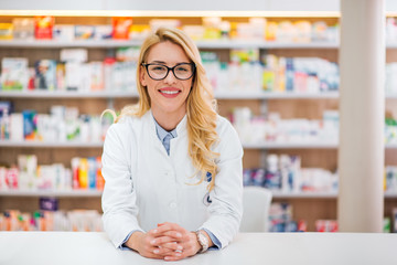 Portrait of a beautiful blonde pharmacist leaning on counter at pharmaceutical store.