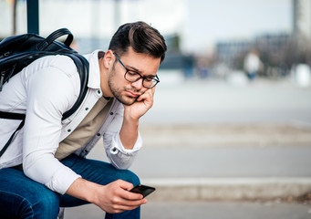 Casual bored young man with backcpack sitting on bench and waiting for bus, using smartphone. - obrazy, fototapety, plakaty