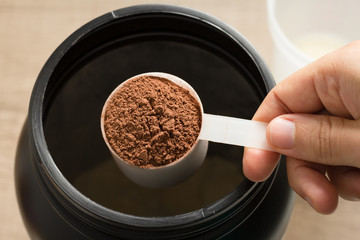 Whey Protein. Point of view of hand holding measuring scoop. Chocolate flavour. Wooden background.