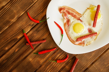 Omelet with bacon, small sausages and red chili peppers in a white plate on a natural wooden surface made of pine boards. Daylight