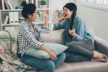 Two laughing asian female friends talking in living room at home sitting relaxing on couch. girls...