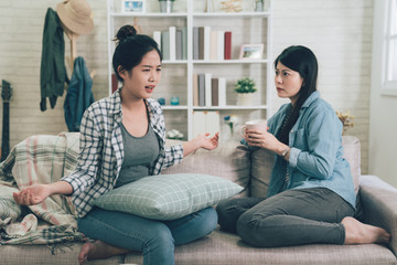 Two female friends sitting on sofa chatting talking and drinking tea or coffee spending time together on holidays. asian woman complain to roommate about incredible thing with hand gesture frowning - obrazy, fototapety, plakaty