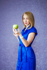 A woman in a blue dress on a purple background holding a green apple and smiling