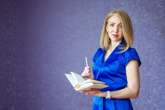 A woman in a blue dress on a purple background holding a white notebook and pen - Powered by Adobe