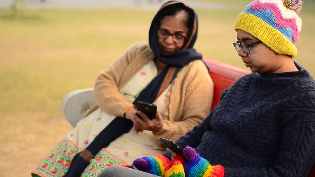 Mother and daughter wearing winter clothes sweater mittens shawl cap and busy working on a mobile pone in an outdoor park 