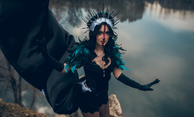 A woman in the image of a fairy and a sorceress standing over a lake in a black dress and a crown.