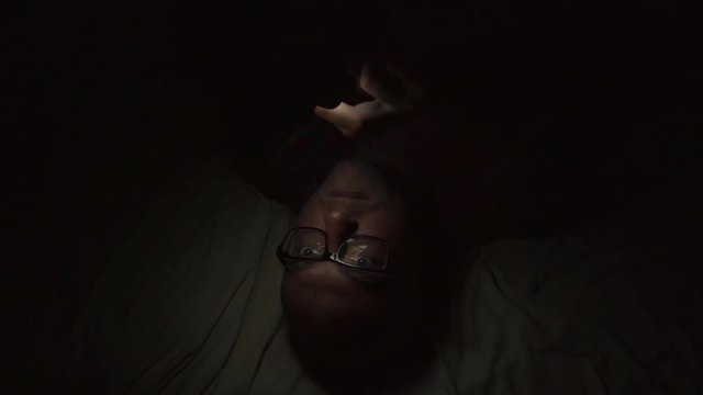 Close-up shot of a guy laying in bed at night and surfing web on smartphone.