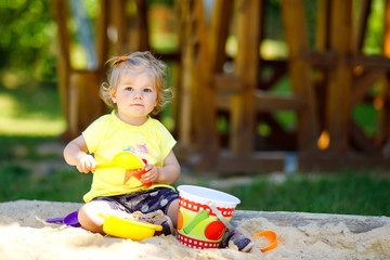 Cute toddler girl playing in sand on outdoor playground. Beautiful baby having fun on sunny warm summer sunny day. Happy healthy child with sand toys and in colorful fashion clothes.