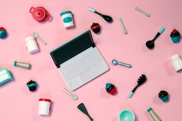 flat lay image of plastic toy tableware with cakes and laptop on pastel pink background