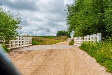 Fototapeta premium Small bridge on a dirt road with white fence next to it some trees and a clouded sky, electricity posts and plantations, seen from a car