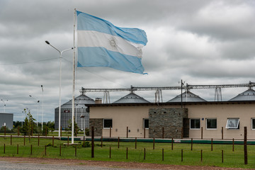 Aged Argentine flag waving in front of an industry with silos on a cloudy day