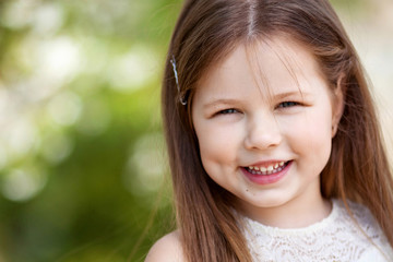 Fototapeta premium Beautiful smiling little girl in cream dress, against green of summer park