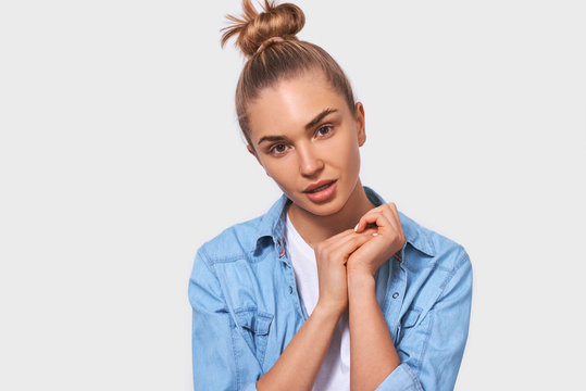 Studio Close Up Portrait Of Adorable European Blonde Young Woman With Bun Hairstyle Dressed In Blue Shirt, Standing Over White Background, Holding Hand With Warm Expression. People Emotions