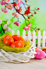 Colorful Easter eggs, white nest and fence on wooden table and branches with spring flowers on green nature background