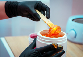 Close-up hands of cosmetologist in black gloves preparing golden colored waxing paste on spatula for sugaring depilation, hair removal beauty procedure.