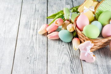 Easter greeting card background with pastel colored eggs and homemade cookies shaped in eggs and bunnies rabbits. With a basket, tulips, rustic wooden table, copy space top view banner