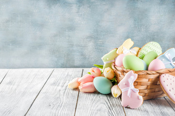 Fototapeta na wymiar Easter greeting card background with pastel colored eggs and homemade cookies shaped in eggs and bunnies rabbits. With a basket, tulips, rustic wooden table, copy space top view banner