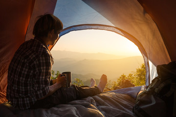 A young man sitting in the tent with holding coffee cup, looking at the mountain landscape in...