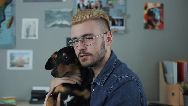 Portrait of caucasian handsome man with glasses and stylish haircut yellow hair sitting at home office desk turn with gog dachshund talking to camera. Speaks motivational speech.