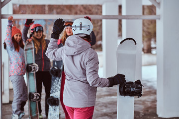 Back view of a woman snowboarder holding a board turned to greet the friends in the winter ski resort