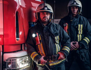 Two firemen wearing protective uniform standing next to a fire engine in a garage of a fire...