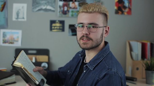 Casual mature hispter man student with stylish haircut yellow hair sitting at home office reading a big brown book. Agains creative wall with beautifull pictures.