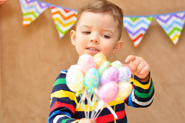 Fototapeta na wymiar Little cute boy in a bright striped raglan smiles and holds a bunch of colorful Easter eggs on sticks