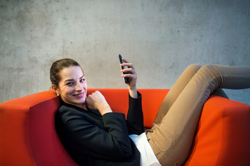 A young businesswoman with smartphone sitting on red armchair in office.