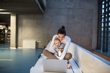 Young student with a baby sitting on desk in room in a library or office, using laptop.