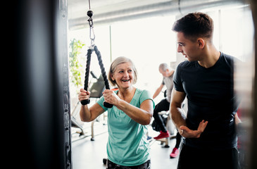 A female senior with a young trainer doing strength workout exercise in gym.