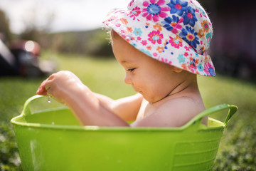 Small girl with a hat in bucket outdoors in garden in summer, playing in water.