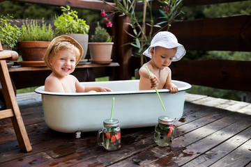 Small children with a drink sitting in bath outdoors in garden in summer.