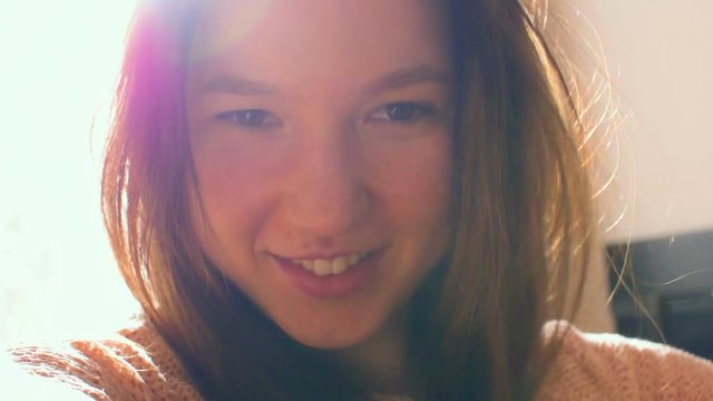 A young woman is looking at the camera and smiling. The light of the sun's rays makes its way out of the window